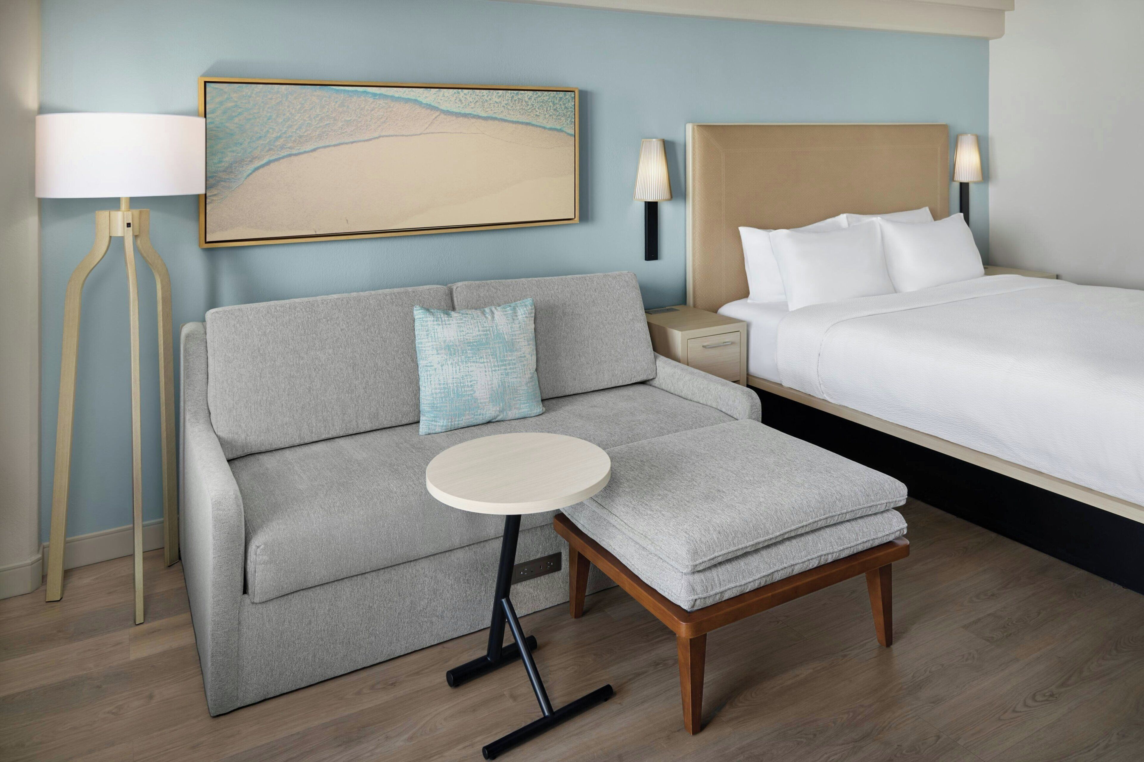 A hotel room with a bed, a sofa, a small table, a lamp, and a wall-mounted artwork.