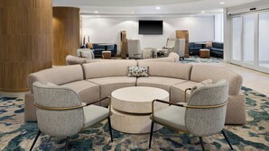 A modern hotel lobby with a curved seating area, a round coffee table, and a flat-screen TV mounted on the wall.