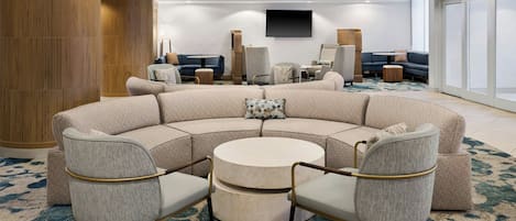 A modern hotel lobby with a curved seating area, a round coffee table, and a flat-screen TV mounted on the wall.