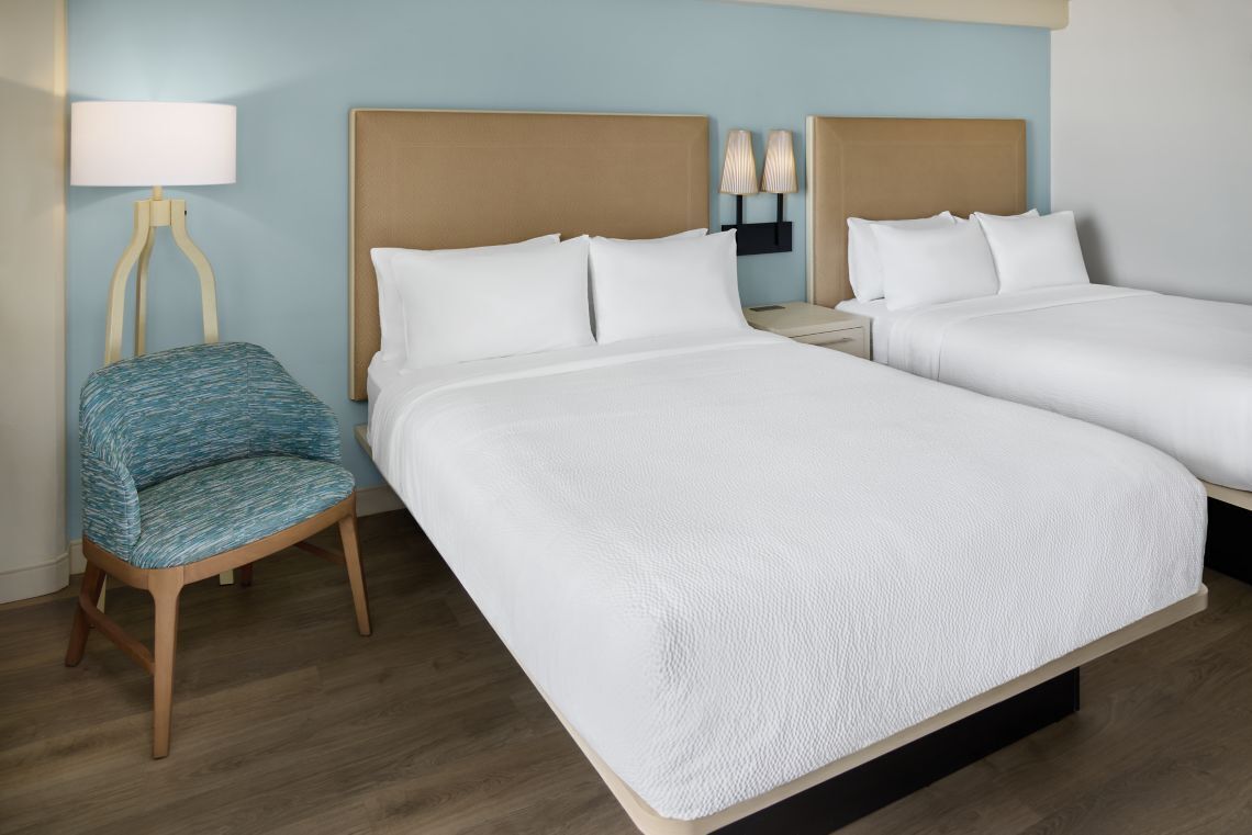 Quarto de hotel com duas camas, uma cadeira, um abajur e uma mesa de cabeceira.