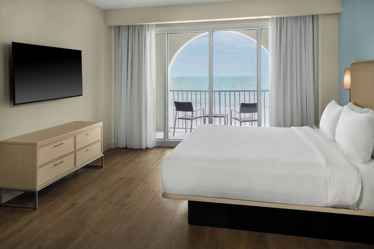 Habitación de hotel con una cama grande, televisor, cómoda de madera y balcón con vistas al mar.