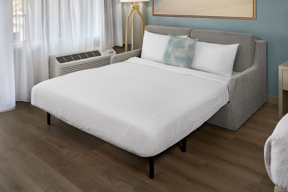 A neatly arranged bedroom with a bed, a sofa, a lamp, and a wall-mounted air conditioner.