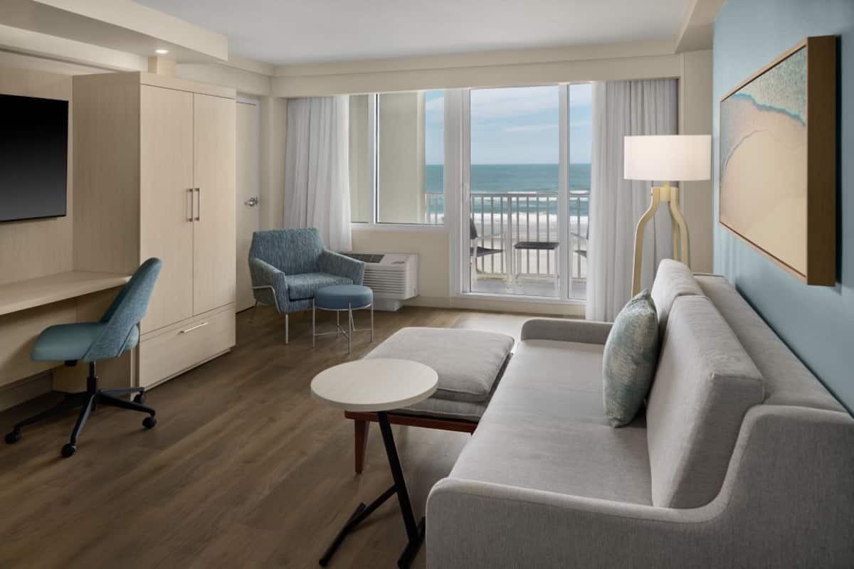 Habitación de hotel con sofá, sillón, mesita, televisión y balcón con vista al océano.