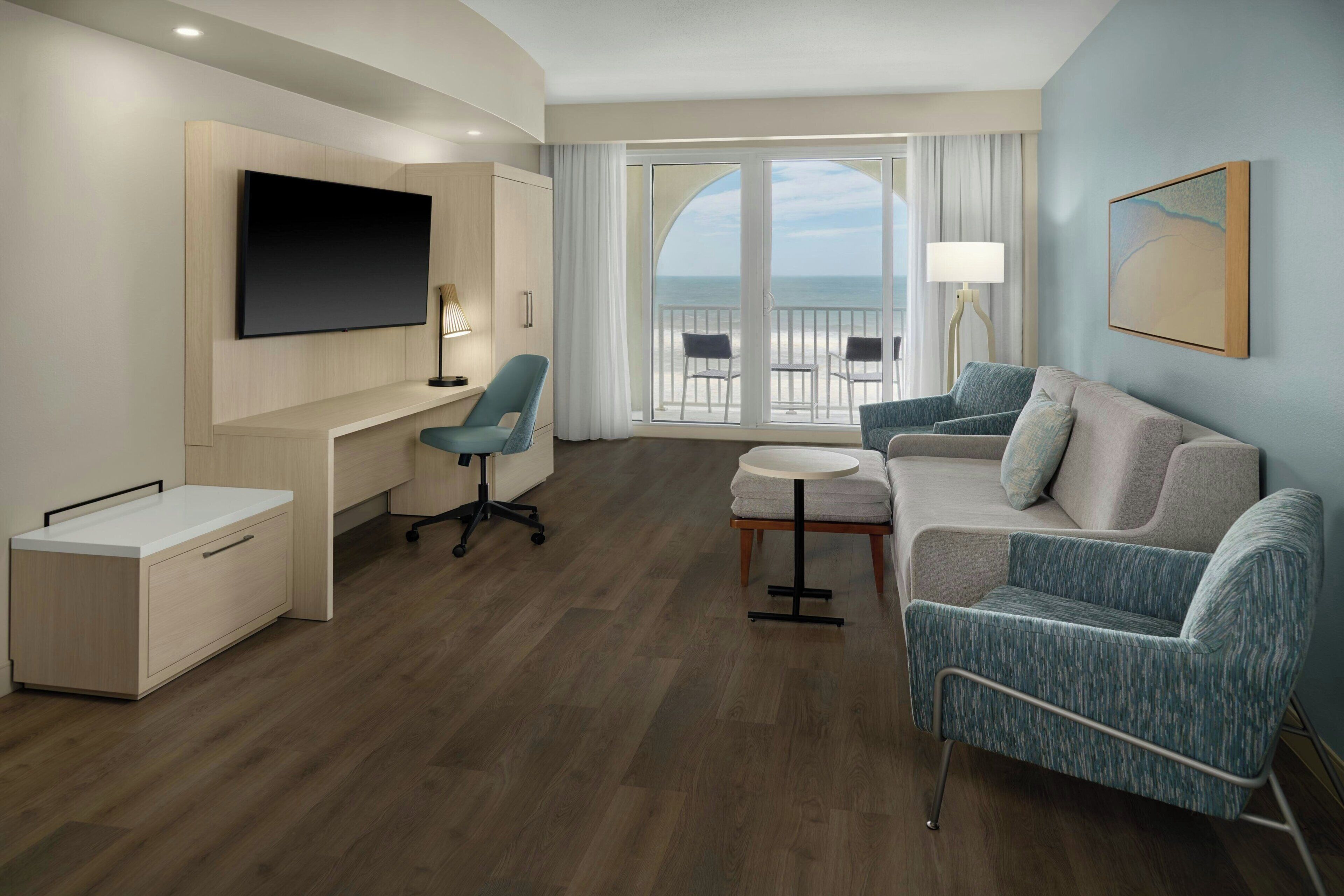 A modern hotel room with a flat-screen TV, a desk, a sofa, and a balcony with a view of the sea.