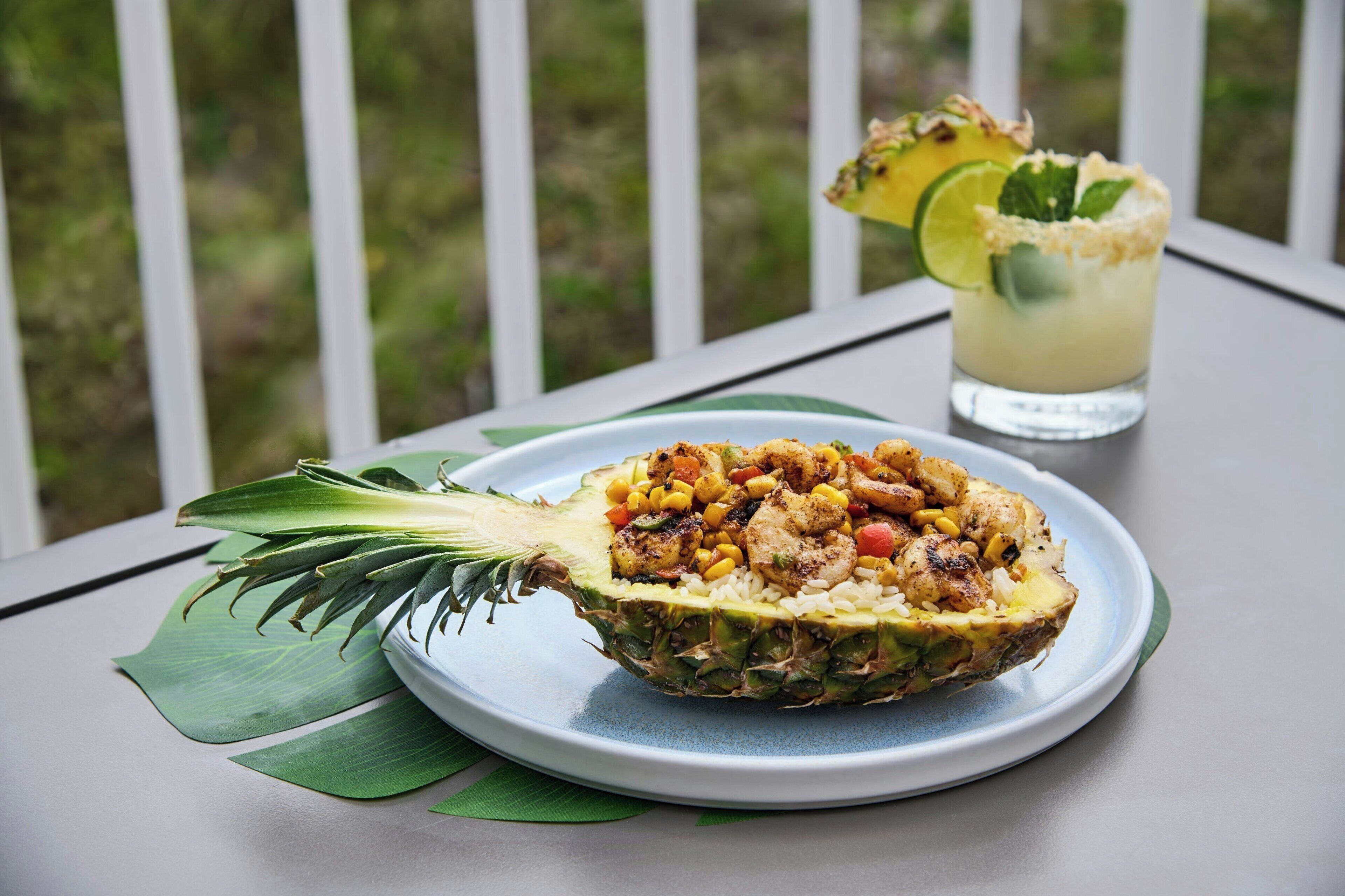 A pineapple bowl filled with a meal, a glass of drink with a lime garnish, and a green leaf on a table.