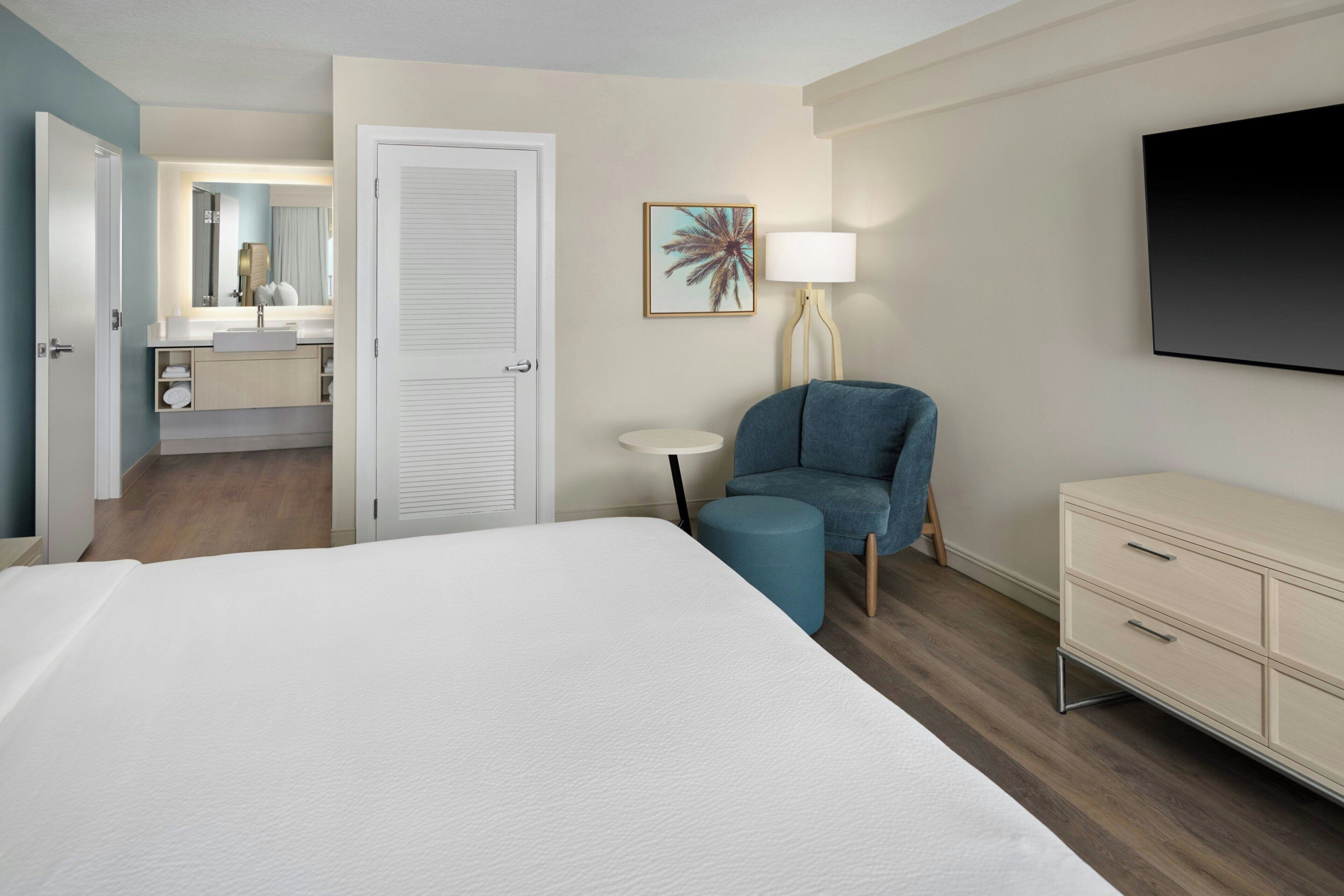 Habitación de hotel con cama, escritorio, silla, televisor, lámpara y baño visible a través de una puerta abierta.