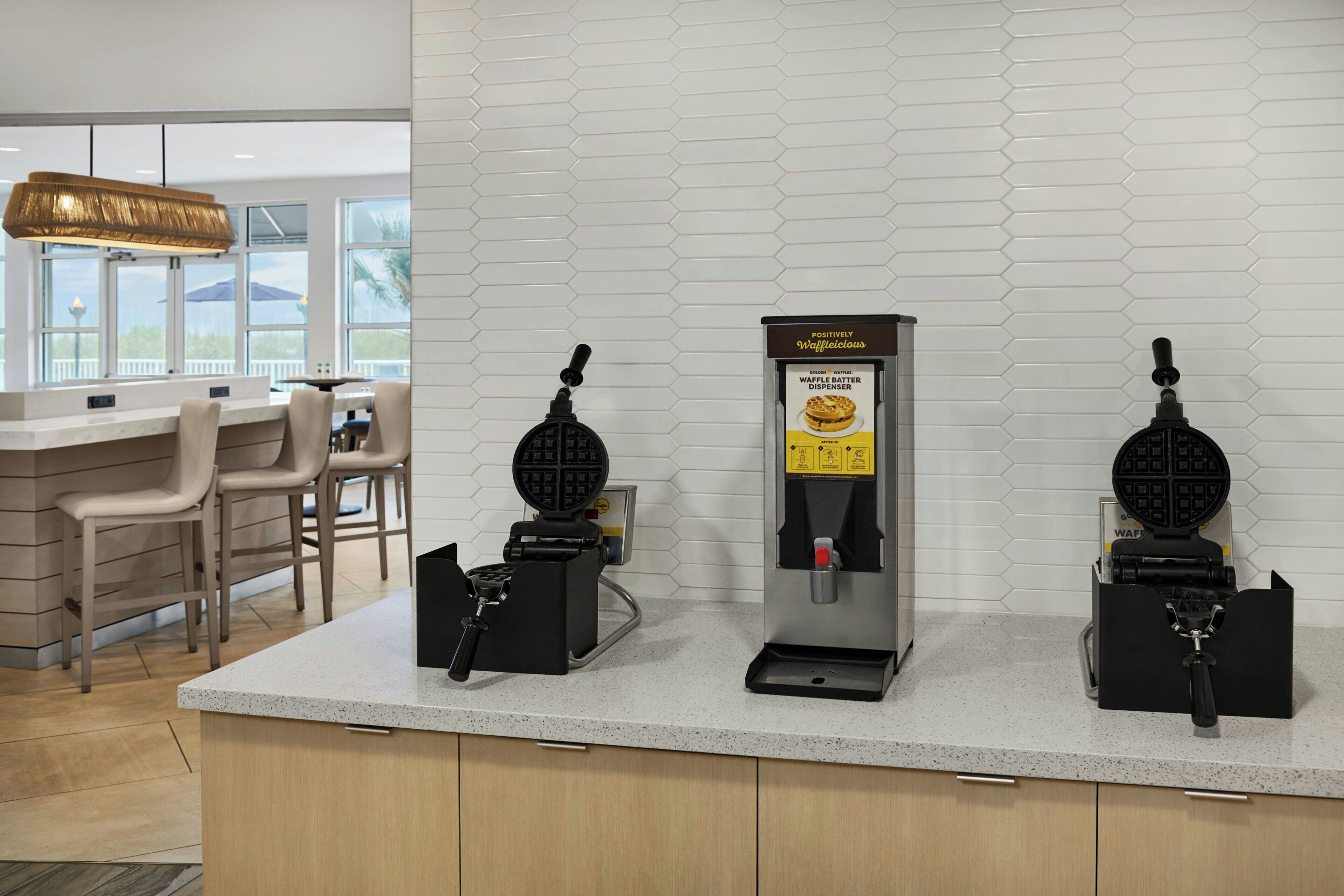 A kitchen area with waffle makers, a waffle dispenser, and a seating area with chairs.