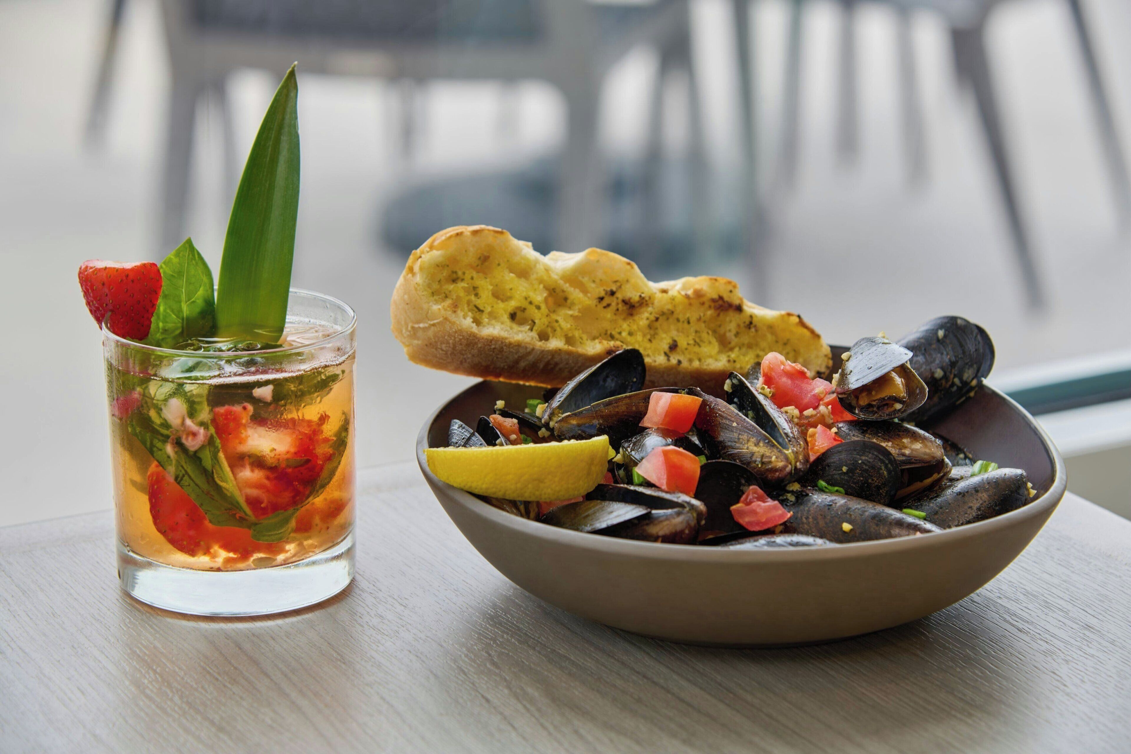 Eine Schale mit Muscheln, Zitrone und Tomaten, ein Glas mit einem Erdbeergetränk und ein Stück Brot auf einem Tisch.