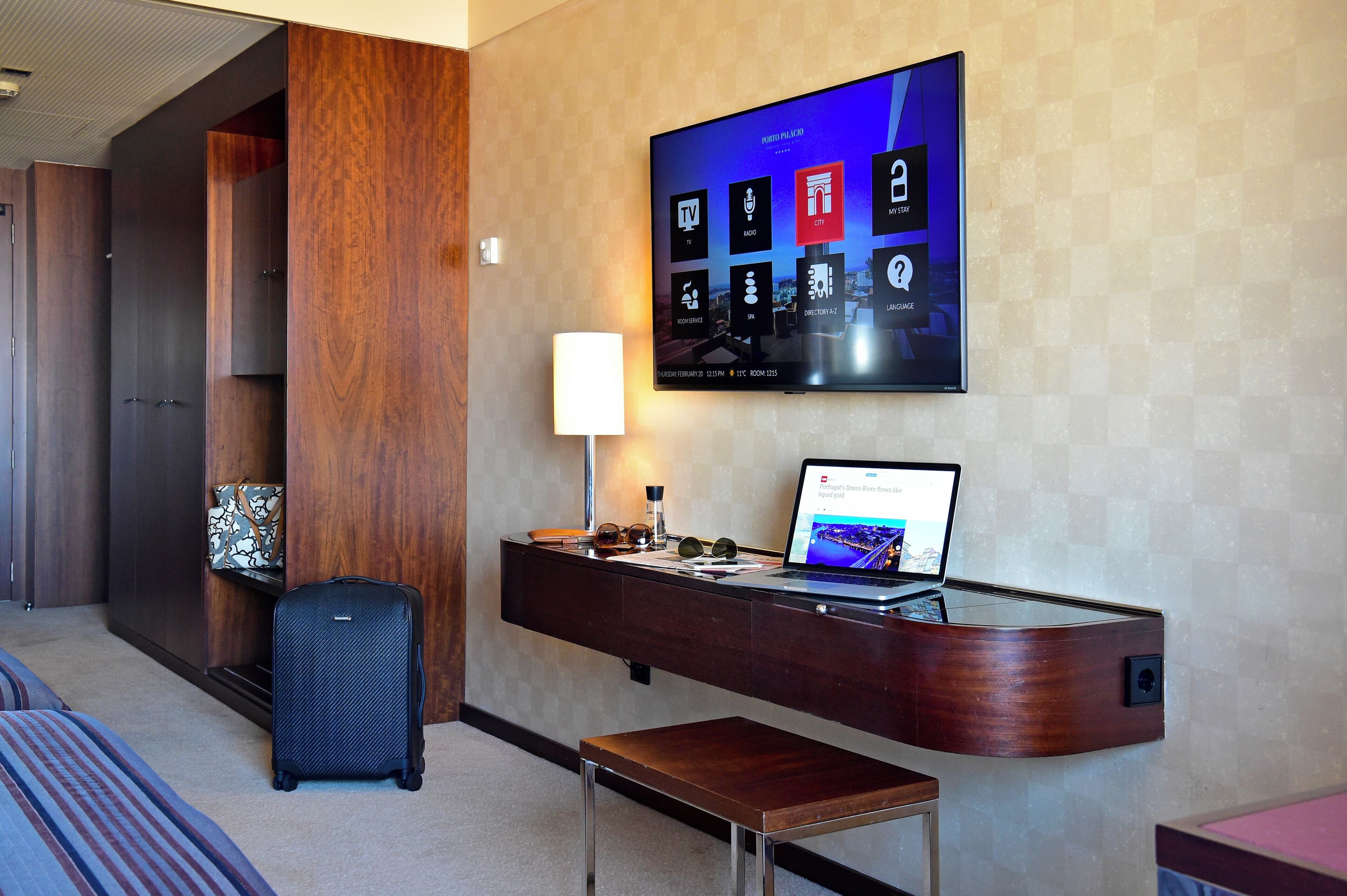 A hotel room with a desk, a laptop, a TV mounted on the wall, a suitcase, and a bench.