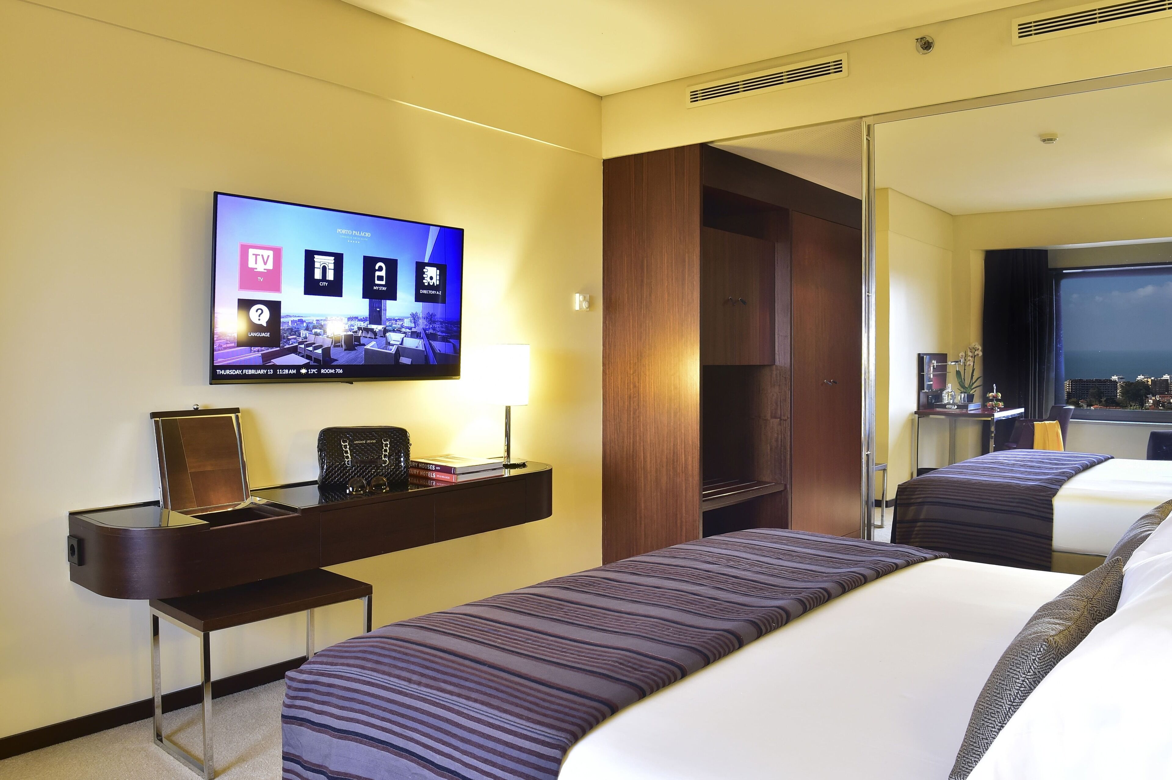 A hotel room with a TV displaying a cityscape, a desk with a lamp, and two beds with striped bedding.