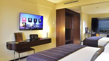 Habitación de hotel con una televisión mostrando un paisaje urbano, un escritorio con lámpara y dos camas con ropa de cama a rayas.