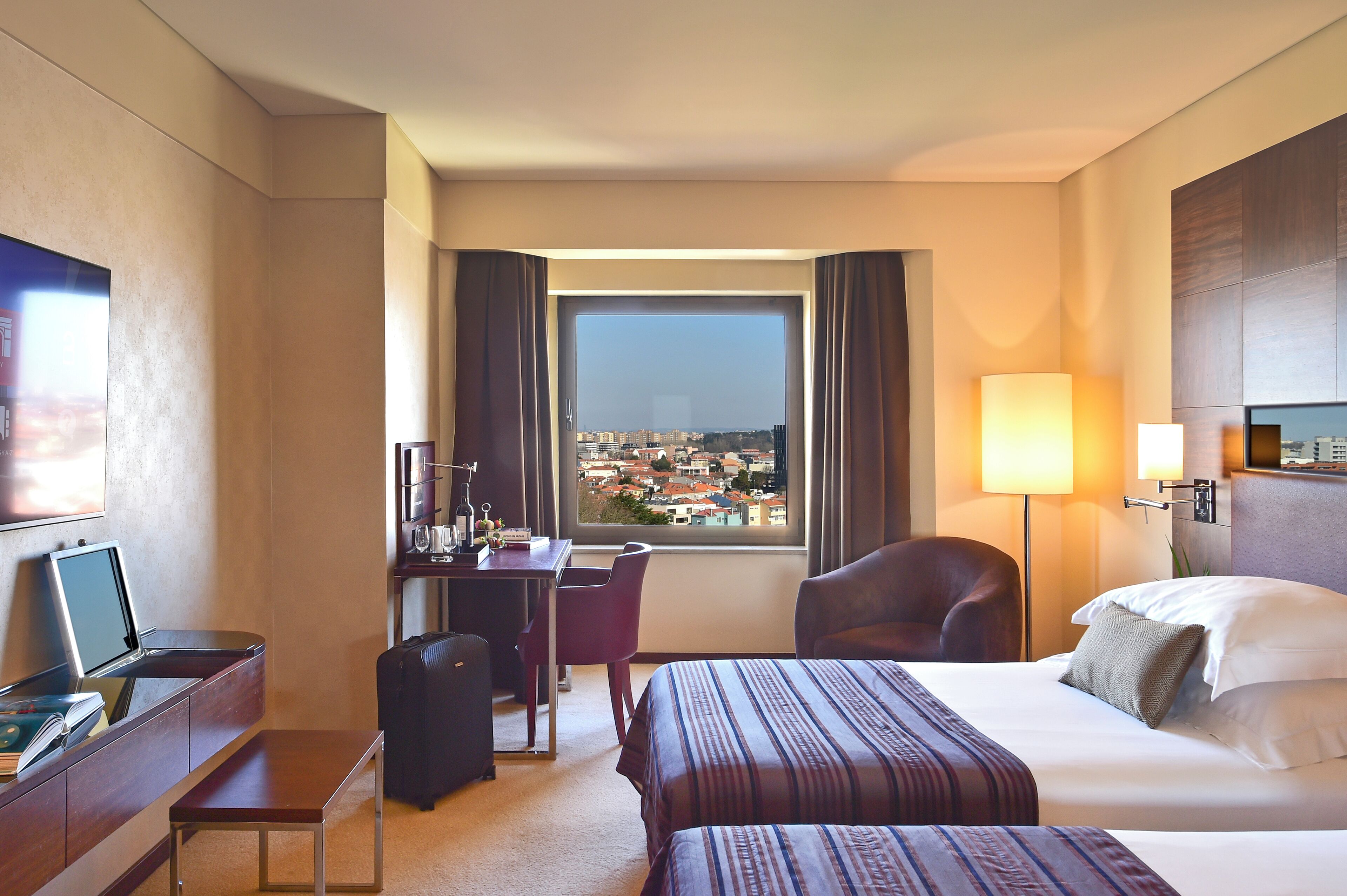Habitación de hotel con cama, escritorio, silla y vistas a la ciudad a través de un ventanal grande.