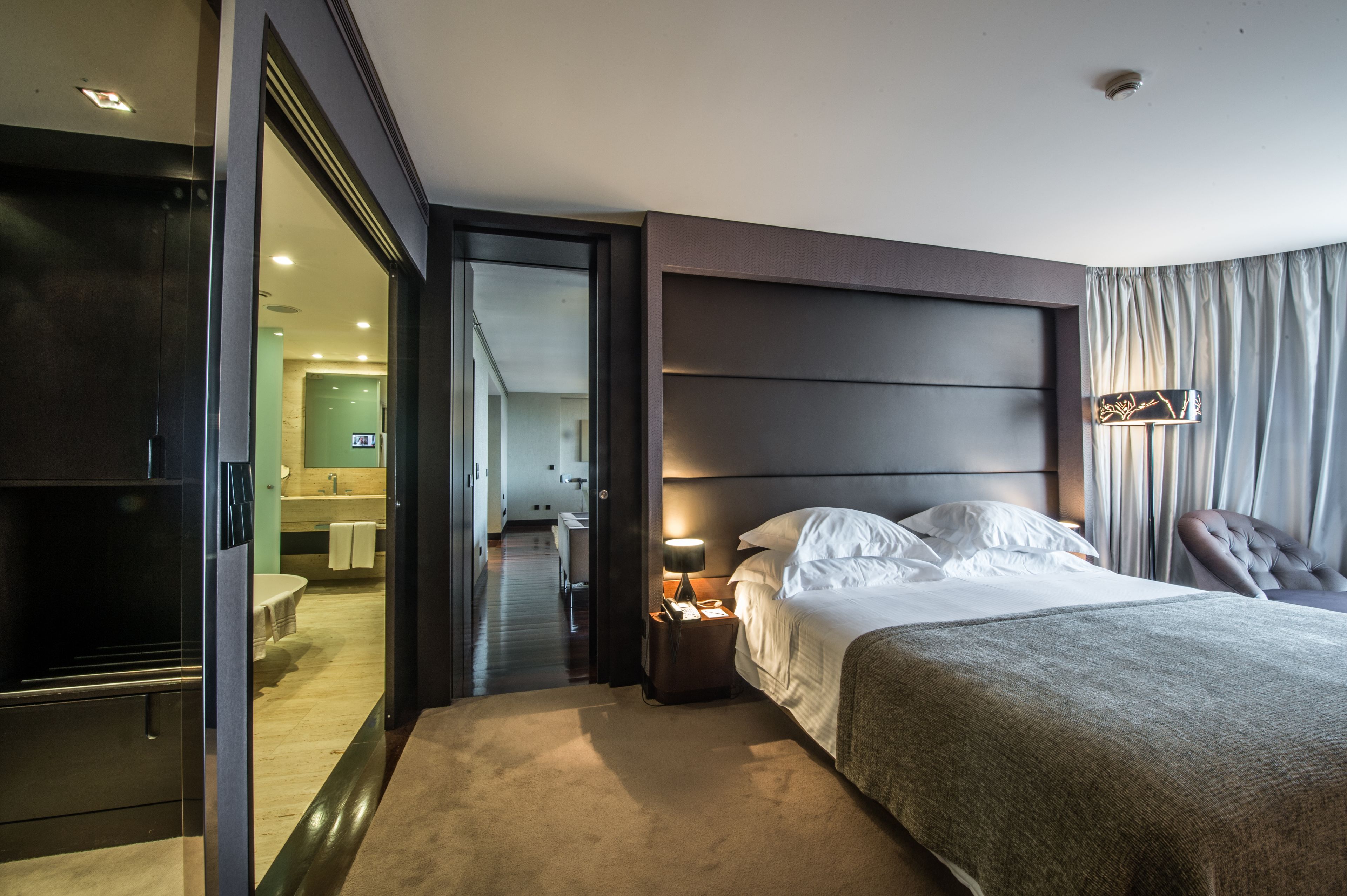 A modern hotel room with a large bed, a nightstand with a lamp, a chair, and a view of the hallway and bathroom.