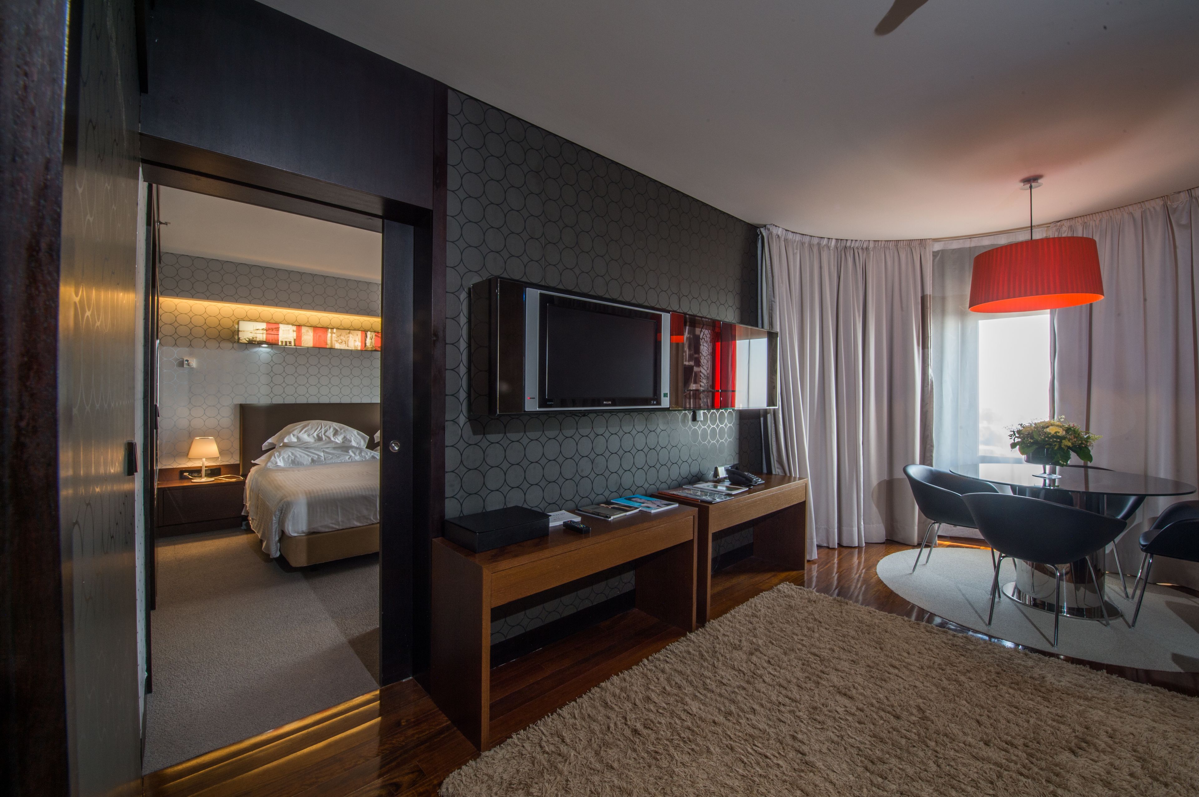 Ein modernes Hotelzimmer mit einem Bett, einem an der Wand montierten Fernseher, einem Holztisch, einer Essecke mit Tisch und Stühlen sowie einer roten Pendelleuchte.