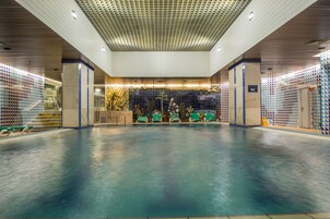 Una piscina interior amplia con suelo de baldosas, sillas verdes y una pared decorativa con un diseño geométrico.