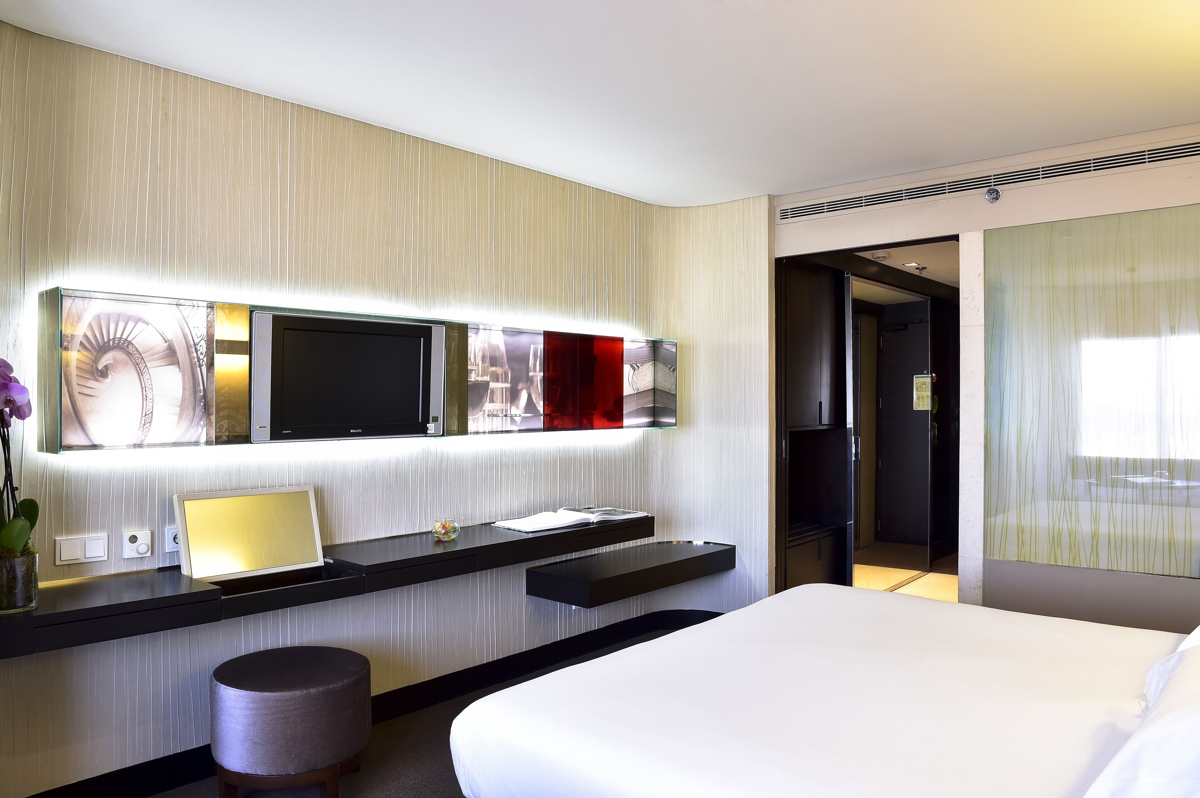 A modern hotel room with a large bed, a built-in TV, a desk with a laptop, and a small stool.