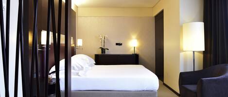A hotel room with a bed, bedside table, lamp, chair, and a decorative plant.