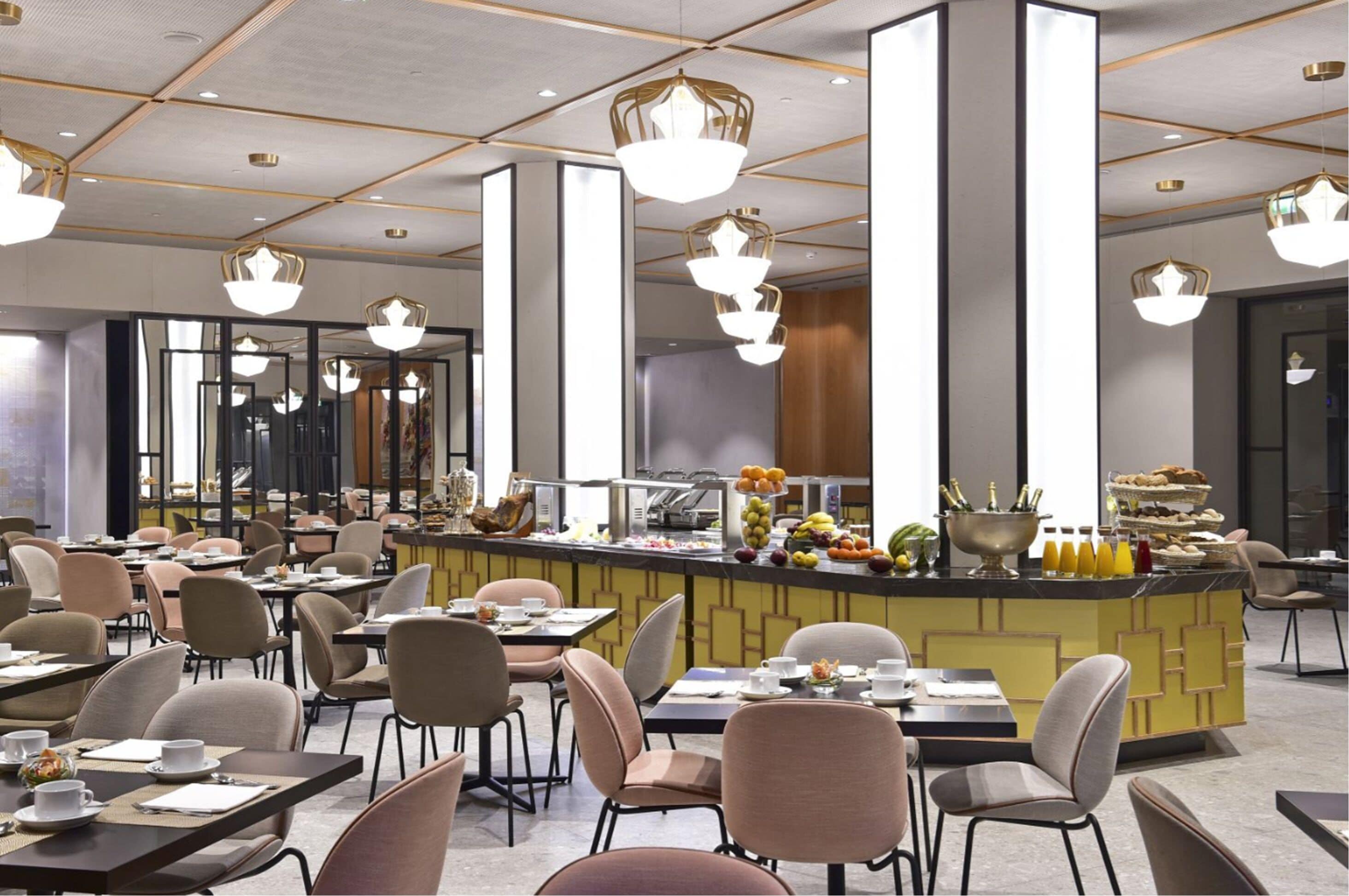 Un comedor moderno con varias mesas y sillas, una barra central con diversas opciones de comida y bebida, e iluminación colgante.