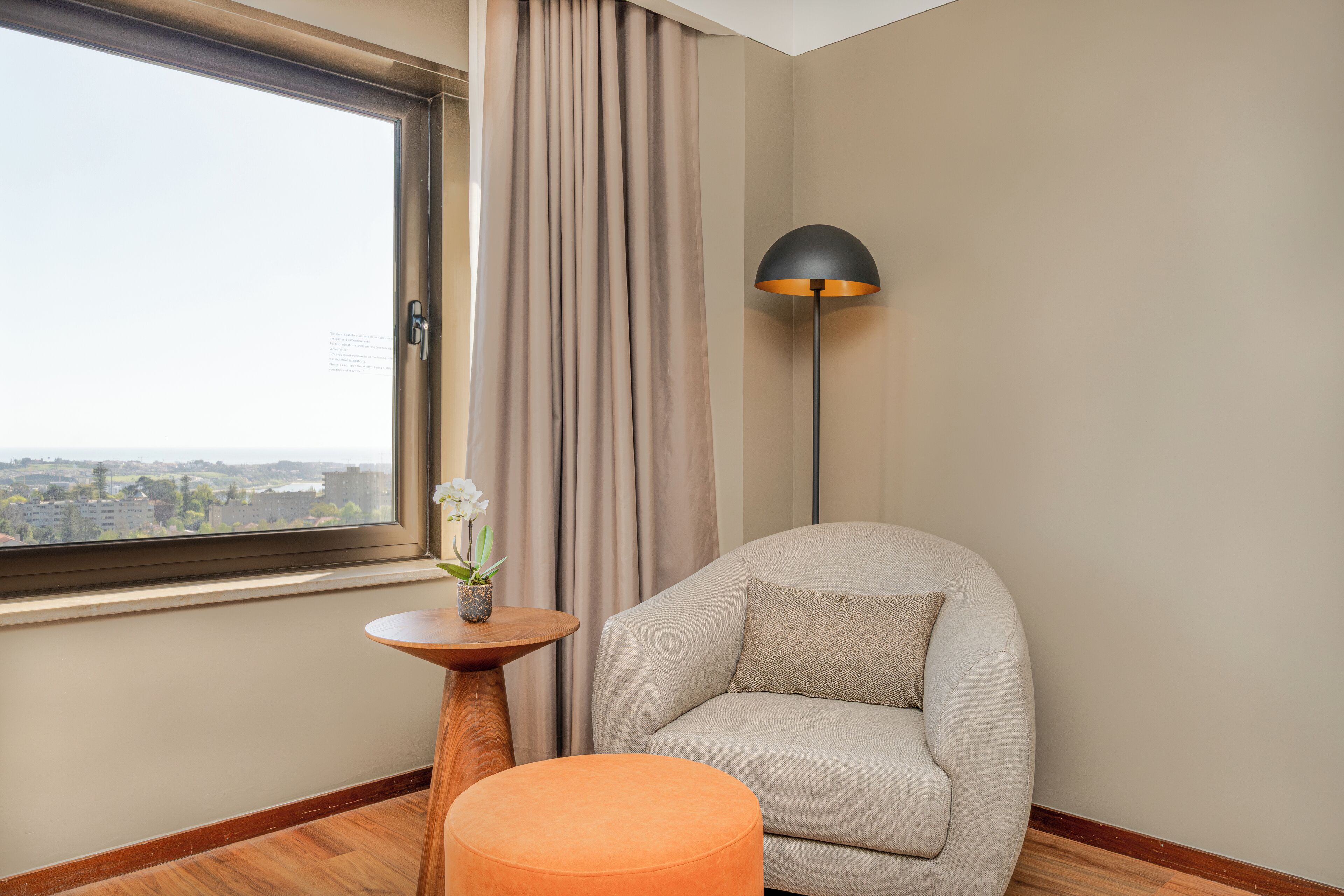 Una sala de estar moderna con un sofá, un otoman naranja redondo, una mesita de madera con un jarrón de flores, una lámpara de pie y vistas de la ciudad a través de un ventanal grande.