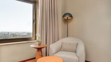 Una sala de estar moderna con un sofá, un otoman naranja redondo, una mesita de madera con un jarrón de flores, una lámpara de pie y vistas de la ciudad a través de un ventanal grande.