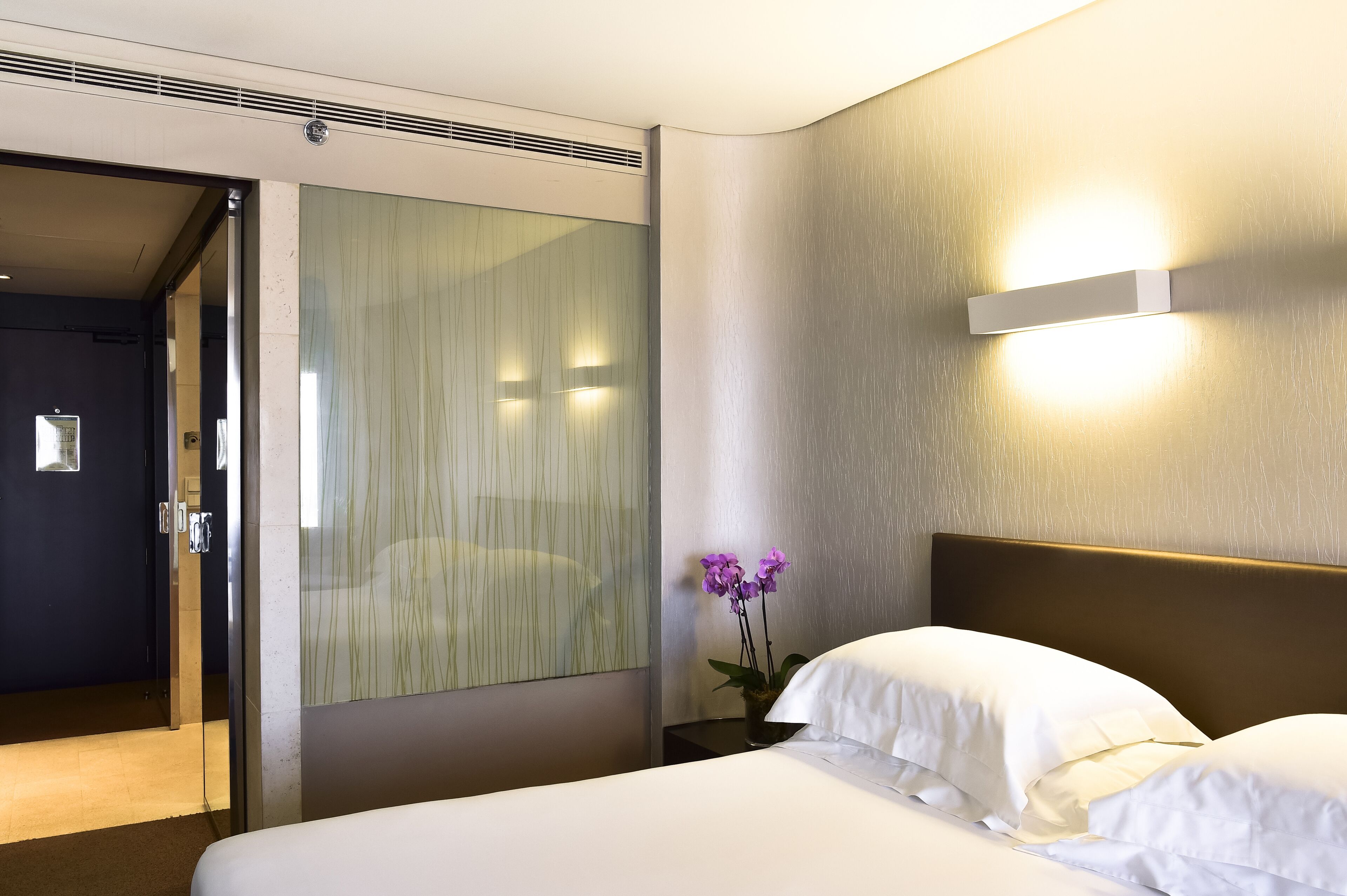 A hotel room with a bed, a nightstand, a vase with flowers, and a wall-mounted light fixture.