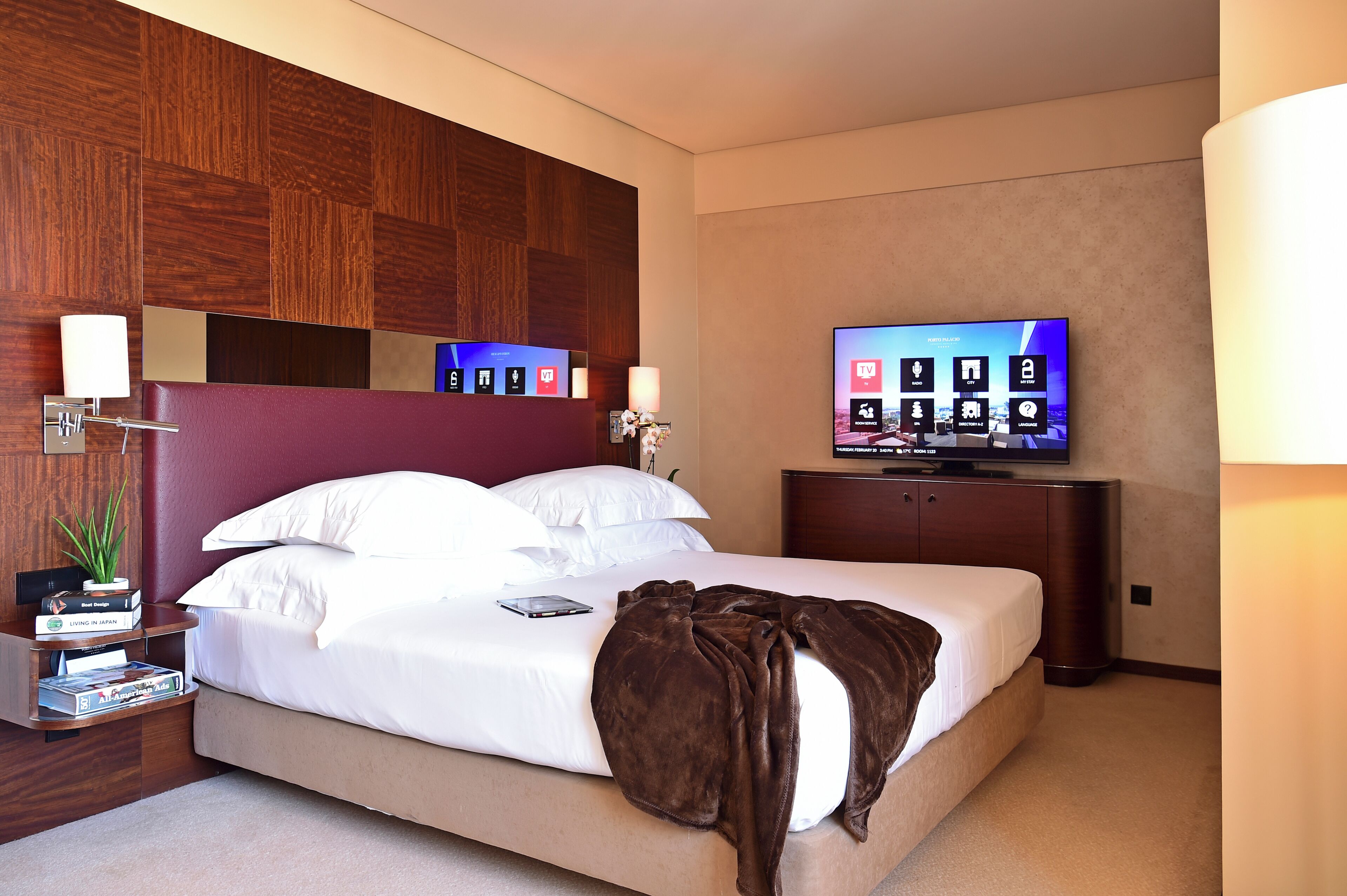 Habitación de hotel con una cama grande, un televisor de pantalla plana, un cabecero de madera y una mesita de noche con una lámpara y libros.