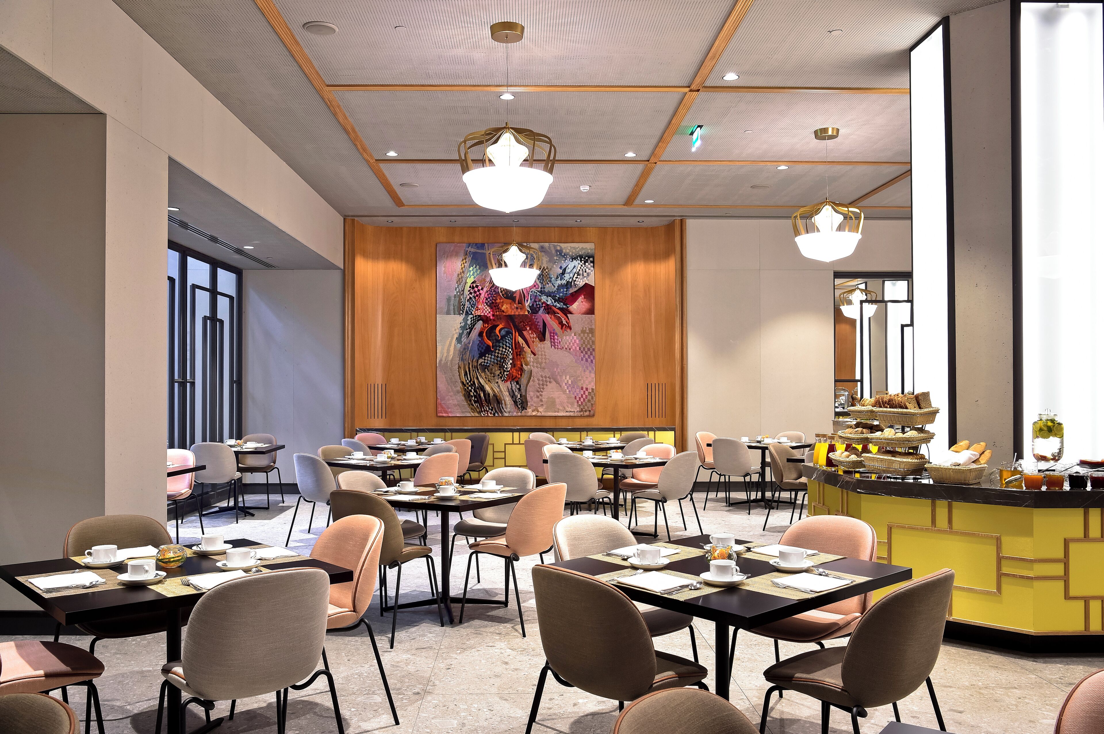 Un comedor de hotel moderno con varias mesas y sillas, una barra central con comida y bebidas, y un gran cuadro abstracto en la pared.