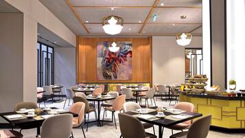 Un comedor de hotel moderno con varias mesas y sillas, una barra central con comida y bebidas, y un gran cuadro abstracto en la pared.