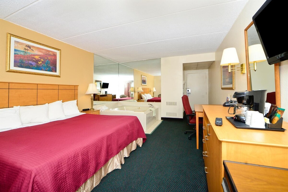 A hotel room with a bed, a desk with a coffee maker, a chair, a mirror, and a television.