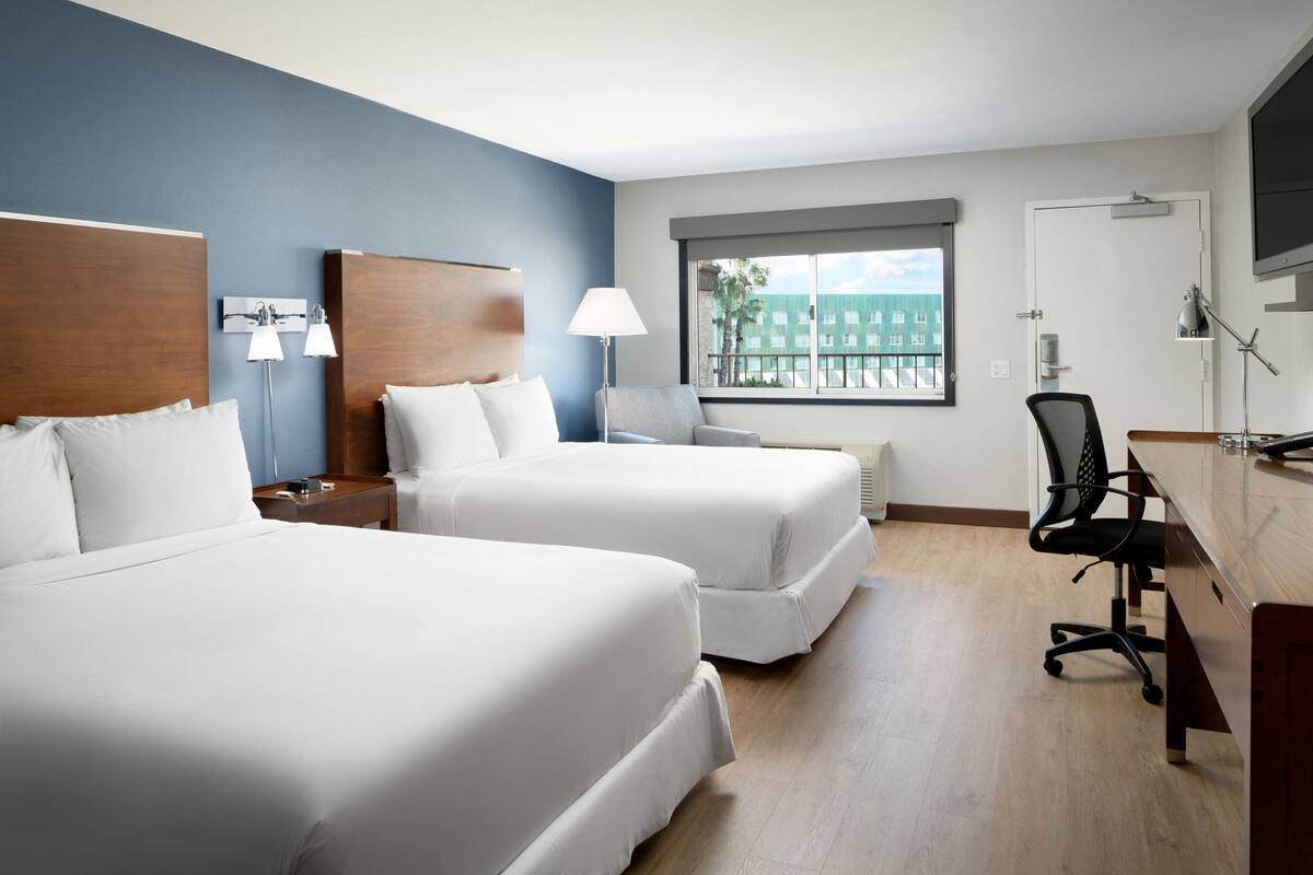 A hotel room with two beds, a desk, a chair, a lamp, and a window with a view of buildings.