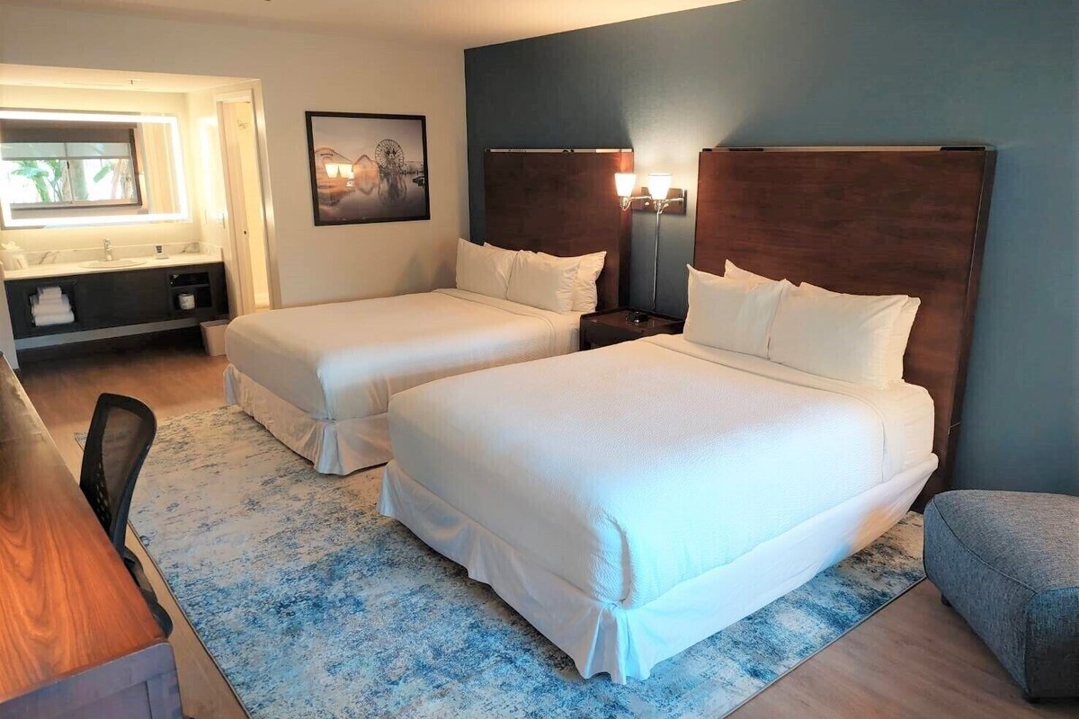 Une chambre d’hôtel avec deux lits, un bureau, une chaise, une table de chevet avec des lampes, un tableau au mur et une fenêtre avec des rideaux.