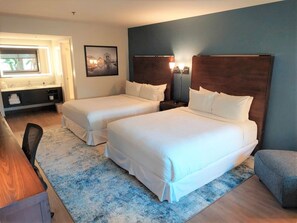Une chambre d’hôtel avec deux lits, un bureau, une chaise, une table de chevet avec des lampes, un tableau au mur et une fenêtre avec des rideaux.