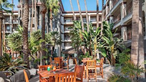 An outdoor patio area with wooden tables and chairs, surrounded by palm trees and a multi-story building with balconies. - Four Points by Sheraton Anaheim (Anaheim)
