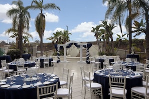 Un salón de bodas al aire libre con mesas redondas, sillas blancas y un arco central.