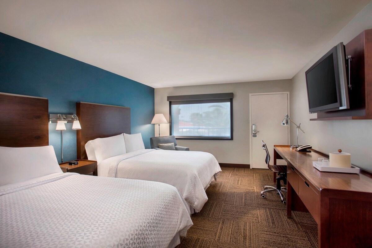 A hotel room with two beds, a desk, a TV, and a window with a view.