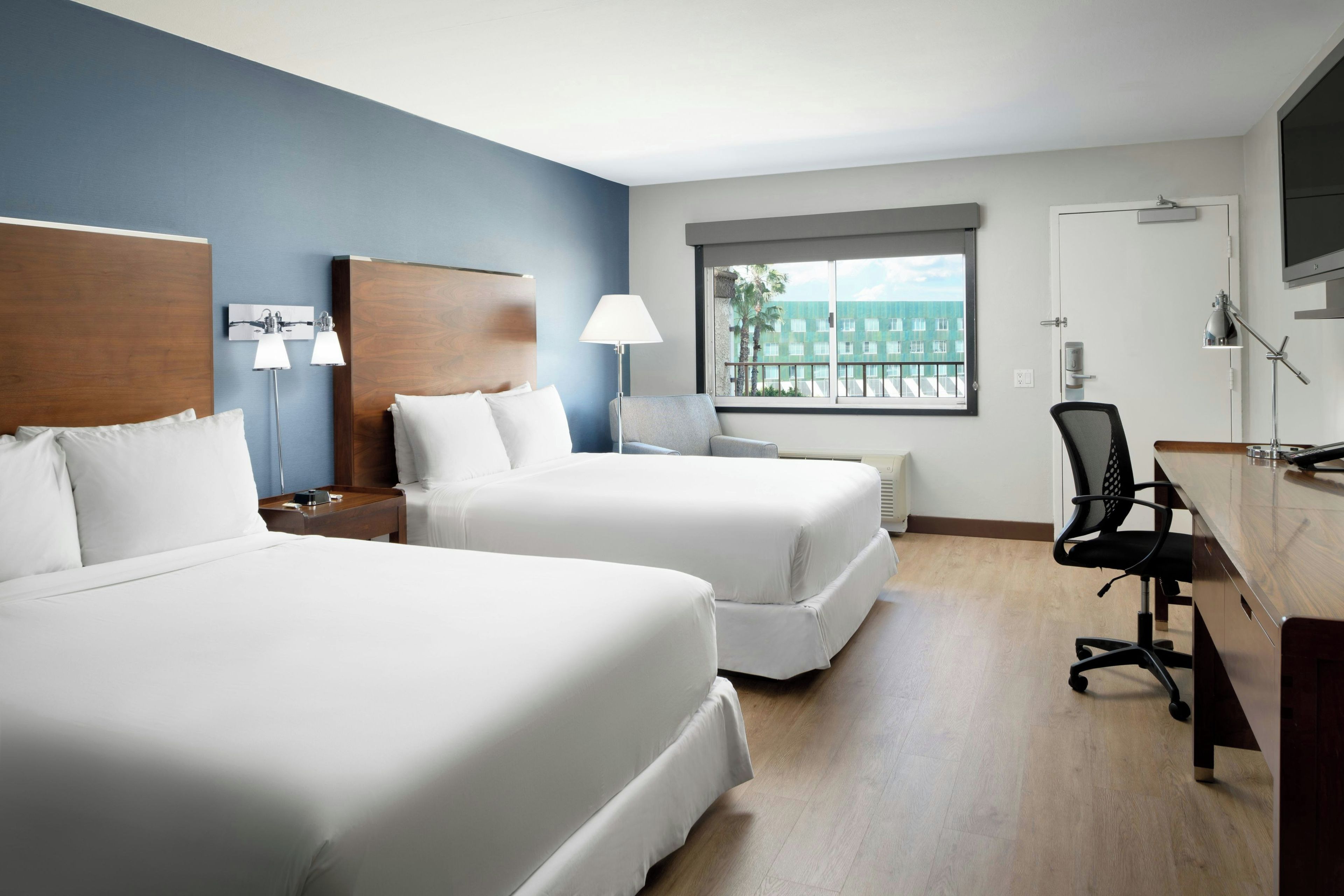 A hotel room with two beds, a desk, a chair, a TV, and a window with a view of buildings.