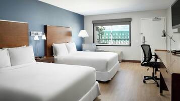A hotel room with two beds, a desk, a chair, a TV, and a window with a view of buildings.