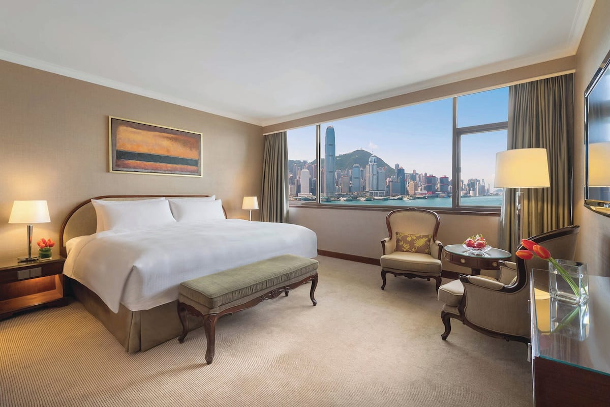 A hotel room with a large bed, a sitting area with a chair and a small table, a view of a cityscape through a large window, and a painting on the wall.