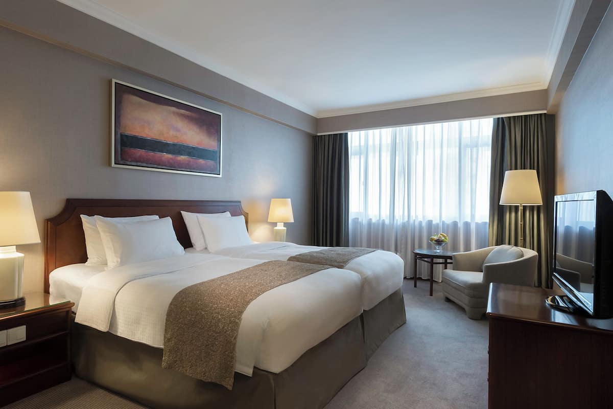 A hotel room with a large bed, two bedside tables with lamps, a TV, a small table with a vase, and a window with curtains.