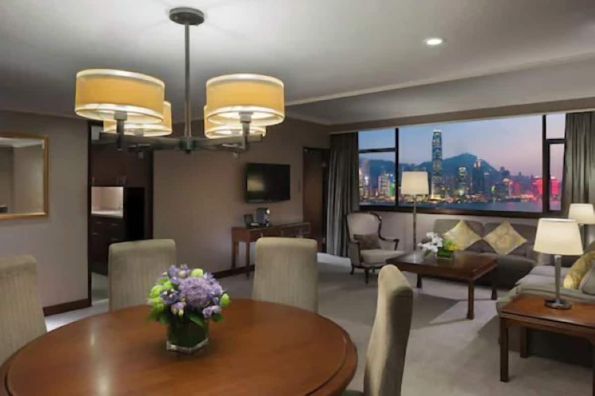 A hotel room with a dining area, a sofa, and a TV. There is a view of a cityscape through large windows.