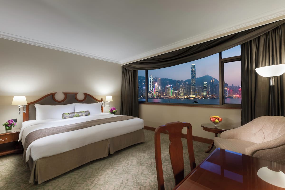 A hotel room with a large bed, a desk, a chair, and a view of a cityscape through a large window.