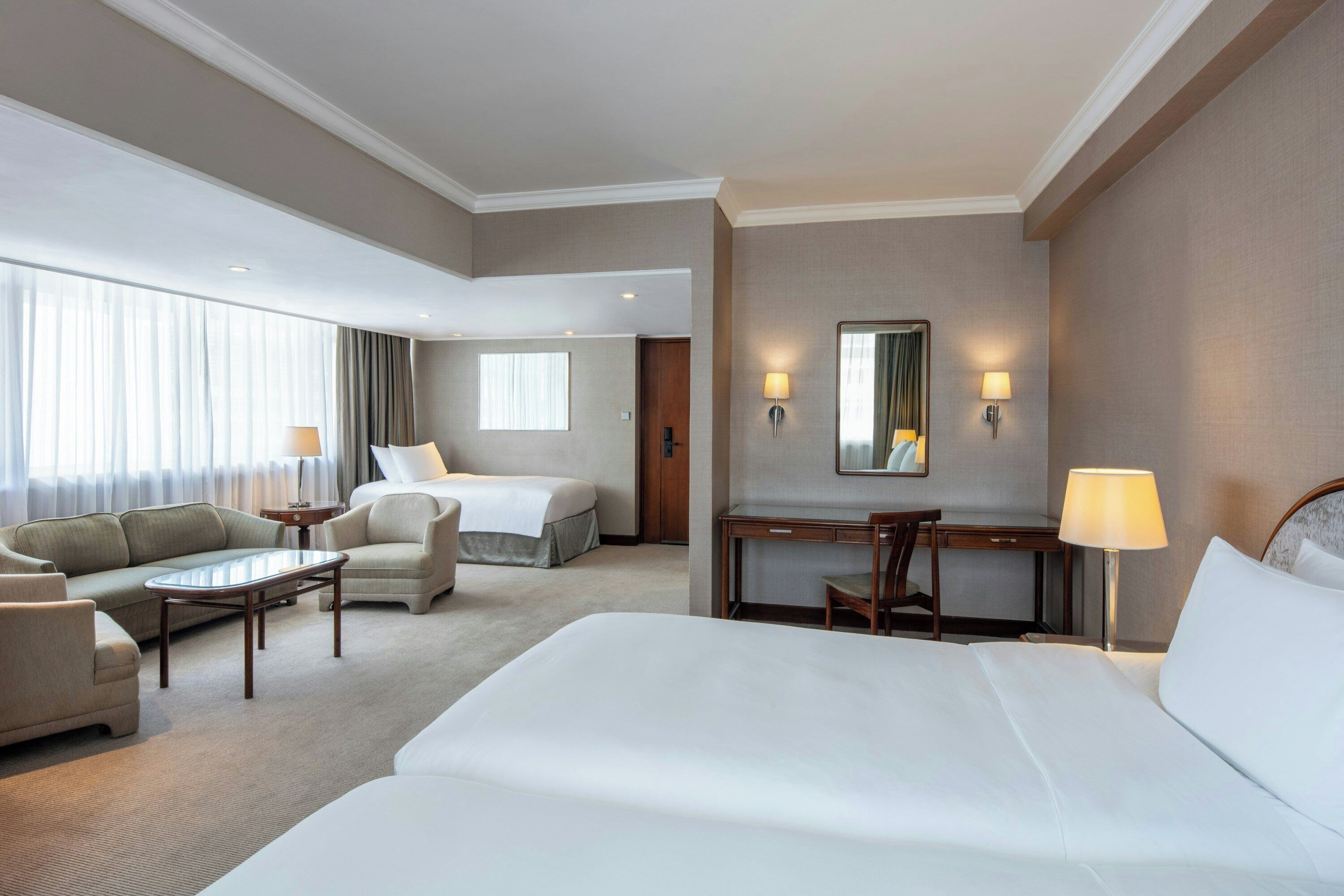 Una habitación de hotel con una cama grande, un sofá, un escritorio y una silla. Hay dos lámparas en las paredes y una ventana con cortinas.