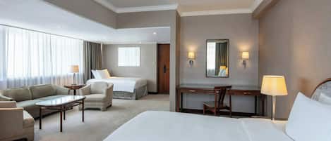 A hotel room with a large bed, a sofa, a desk, and a chair. There are two lamps on the walls and a window with curtains.