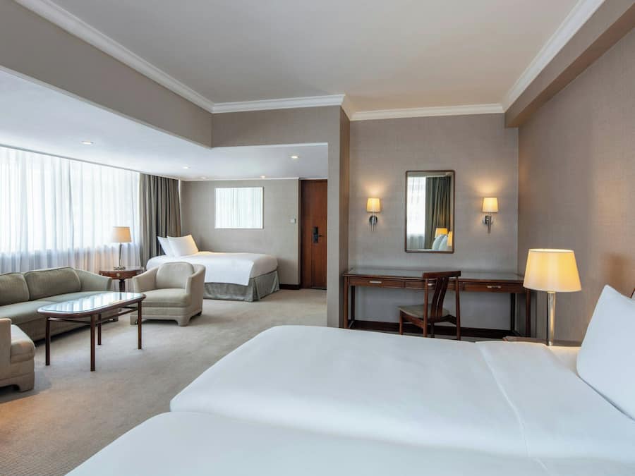 Ein Hotelzimmer mit einem grossen Bett, einem Schlafsofa, einem Schreibtisch und einem Stuhl. An den Wänden befinden sich zwei Lampen und ein Fenster mit Vorhängen.