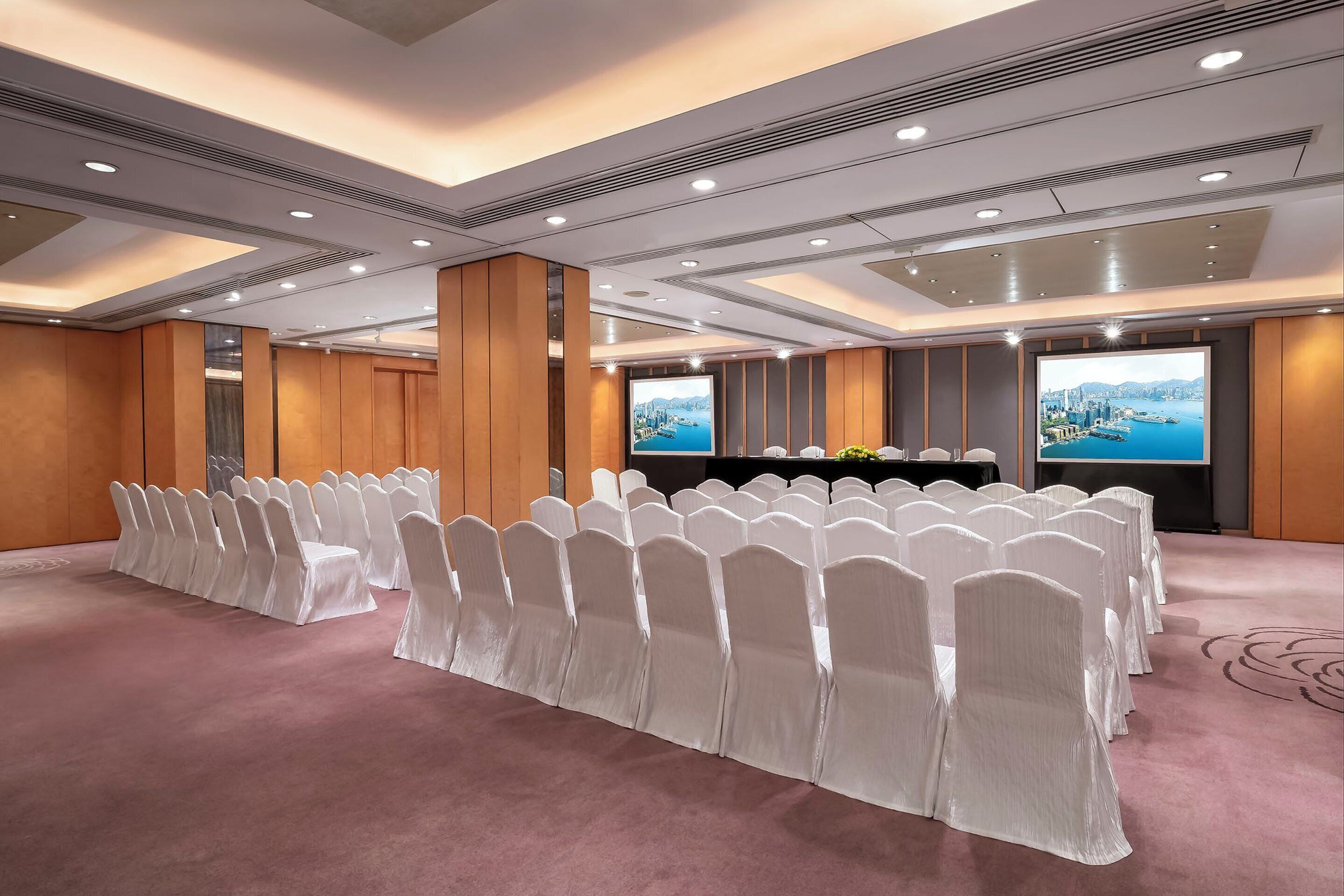 Un salón de conferencias con filas de sillas blancas, una pantalla grande y vistas a un paisaje urbano.