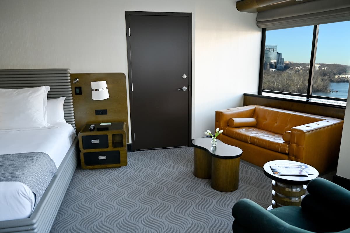 Une chambre d’hôtel avec un lit, un canapé en cuir marron, une petite table avec un vase et une vue sur le paysage urbain à travers de grandes fenêtres.