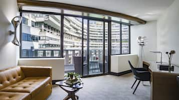 Una sala de estar moderna con un sofá de cuero, una pequeña mesa de centro con una planta, una silla negra y un escritorio con una lámpara. Amplias ventanas ofrecen vistas a rascacielos.