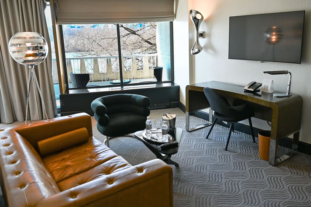 Une chambre d’hôtel moderne, dotée d’un canapé en cuir, d’un bureau avec une chaise, d’une télévision à écran plat et d’une grande fenêtre donnant sur la verdure.