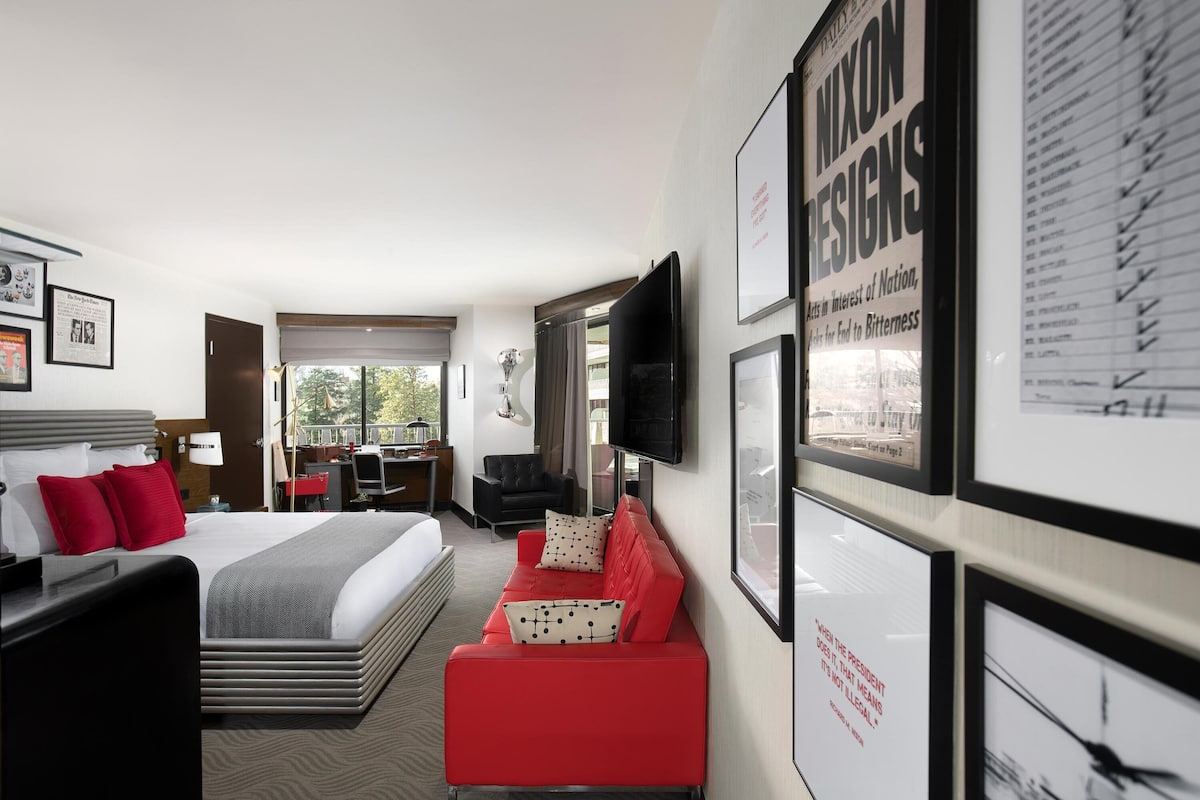 Une chambre d’hôtel avec un lit, un canapé rouge et noir, une télévision et des tableaux encadrés au mur.