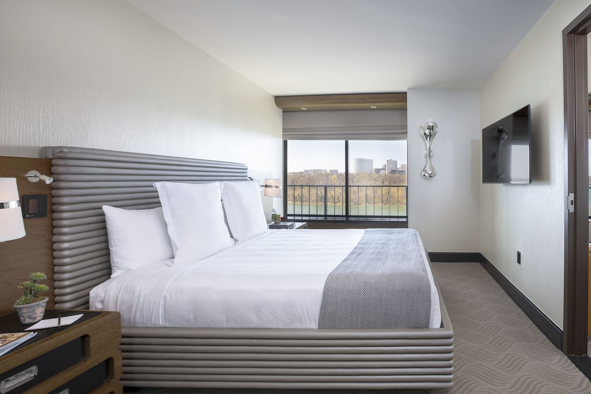 Une chambre d’hôtel moderne avec un grand lit, une table de chevet avec une lampe, un téléviseur fixé au mur et une vue sur la ville par la fenêtre.